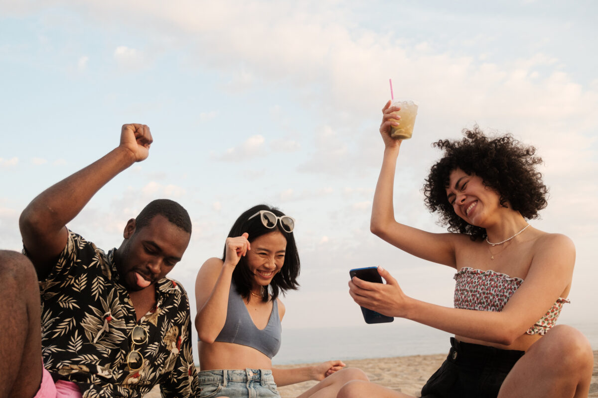 Multi-ethnic young friends sitting on beach towel, drinking some beverages and dancing while listening music with a phone a summer day